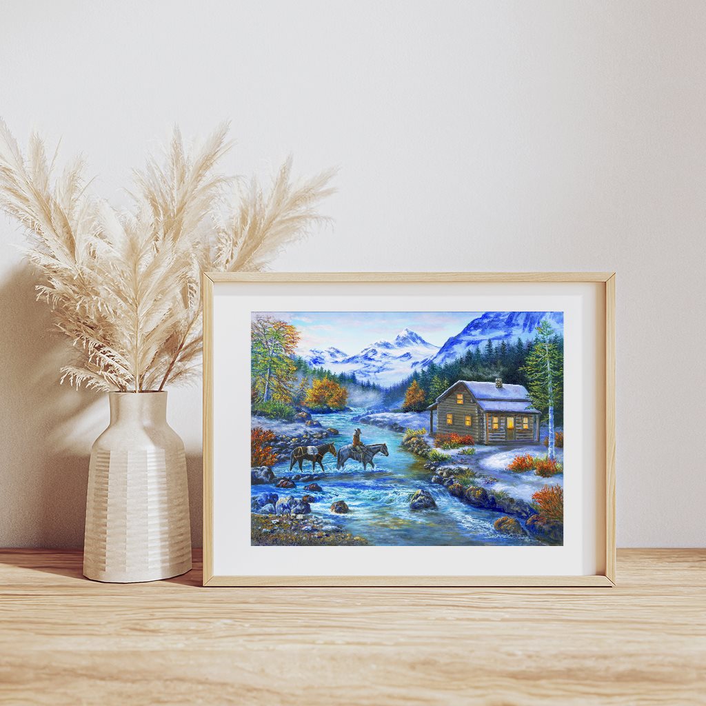 "Home for Supper" Giclée Paper Art Print in natural wood frame, resting on wooden table - Homesteading, cozy log cabin winter scene with horse and pack mule by artist Jeanne Warren