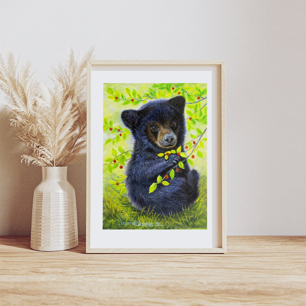 Huckleberry Bear - Black bear cub giclée paper art print in natural wood frame, resting on wood table, by artist Jeanne Warren