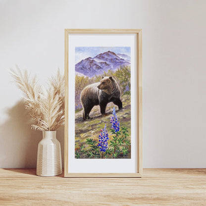 Grizzly & Lupine paper print in a natural wood frame, resting on a wood tabletop. By Artist Jeanne Warren